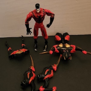 May include: A red and black action figure with a black mask and a spider symbol on its chest. The figure is standing on a white surface. There are several other pieces of the figure, including a pair of legs, a torso, and a head. The pieces are all black and red.