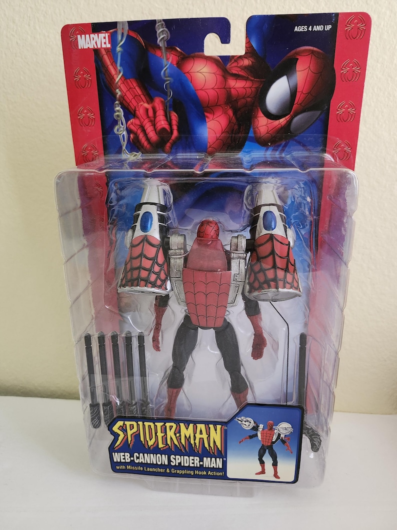 May include: A red and black action figure of Spiderman wearing a web-cannon suit. The figure is packaged in a plastic blister pack with a blue and red background. The packaging includes the text "Spiderman Web-Cannon Spiderman with Missile Launcher & Grappling Hook Action!"