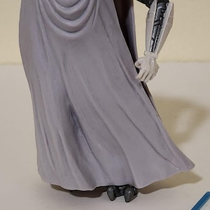 Może przedstawiać: Figurka akcji Generała Grievousa z jasnoszarym płaszczem, białą głową i czarno-srebrnymi mechanicznymi ramionami. Figurka pozuje z blasterem i niebieskim mieczem świetlnym. Przedmiot kolekcjonerski.