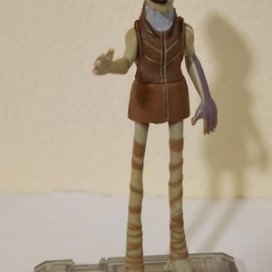 Könnte beinhalten: Detaillierte Actionfigur eines humanoiden Alien-Charakters. Die Figur hat ein braunes und beiges Farbschema mit einer braunen Weste und einem Rock. Die Figur steht auf einer transparenten rechteckigen Basis. Die Figur hat lange, dünne Beine und Arme.