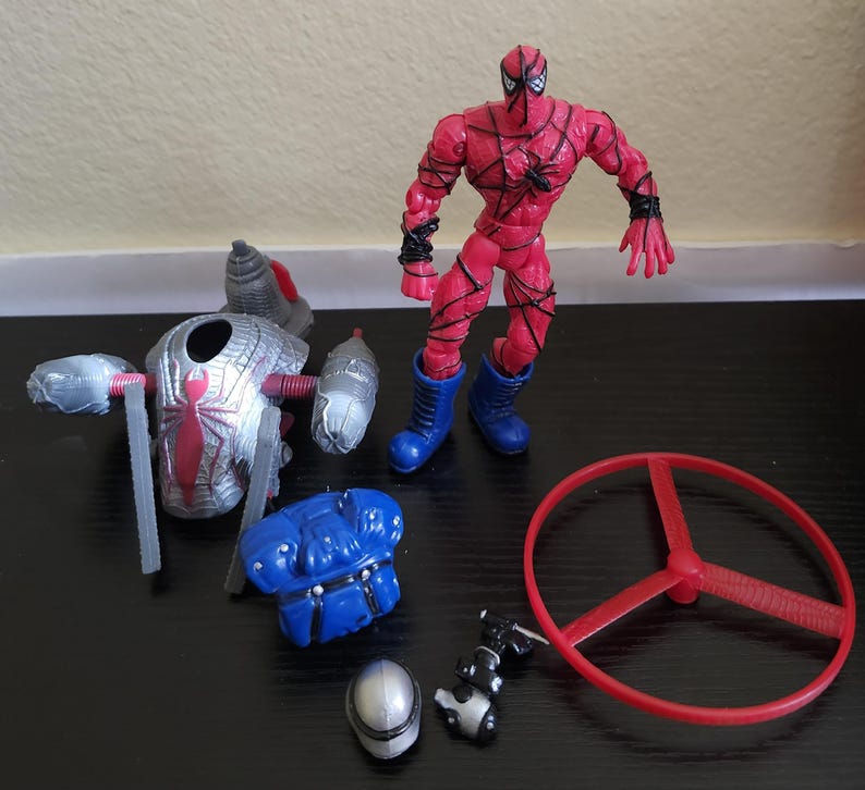 May include: A red and black Spider-Man action figure with blue boots, alongside accessories. Accessories include a silver and red spider-themed device, a blue chest piece, a red propeller, a silver helmet, and a black and silver weapon.