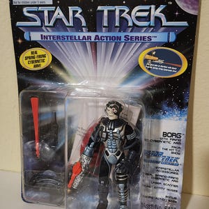 May include: A Star Trek Interstellar Action Series Borg action figure in its original packaging. The figure is wearing a black and silver uniform and has a red cybernetic arm. The packaging includes the text "Real Spring-Firing Cybernetic Arm!"
