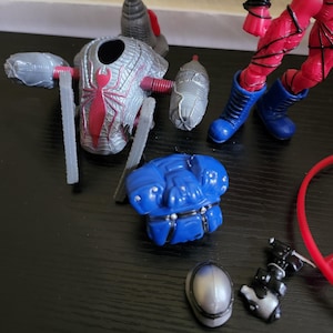 May include: A disassembled action figure with various parts scattered on a dark surface. The parts include a silver and red spider-themed chest piece, blue boots, a blue armored piece, and other accessories. The figure's red legs are also visible.