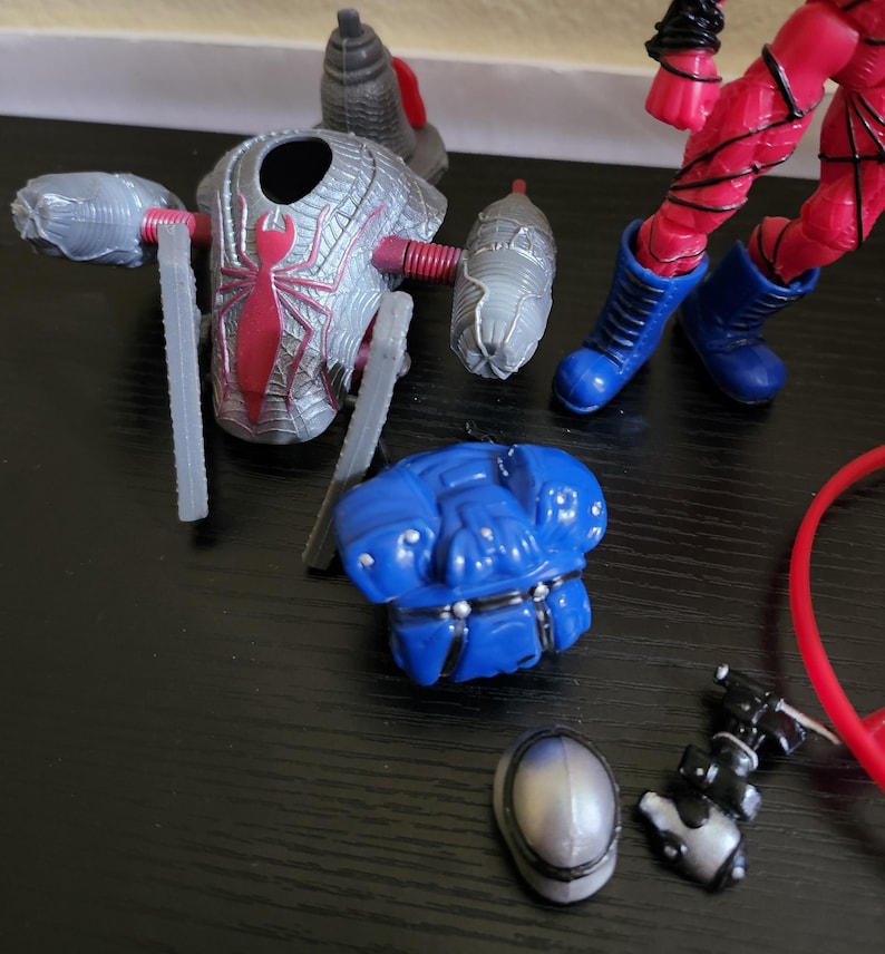 May include: A disassembled action figure with various parts scattered on a dark surface. The parts include a silver and red spider-themed chest piece, blue boots, a blue armored piece, and other accessories. The figure's red legs are also visible.