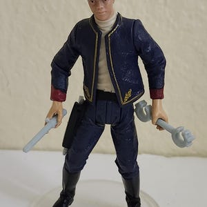 May include: A detailed figurine of a person in a dark blue uniform with gold trim, holding two gray weapons. The figure wears a matching hat and black boots. The base is clear plastic.
