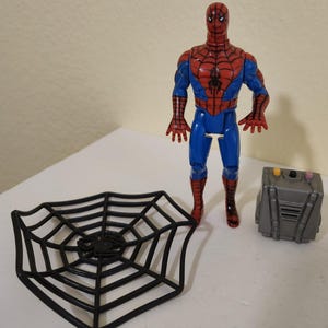 May include: A Spider-Man action figure in a classic red and blue suit, standing next to a black web-shaped accessory and a gray box. The figure has a detailed design, capturing the iconic superhero look.