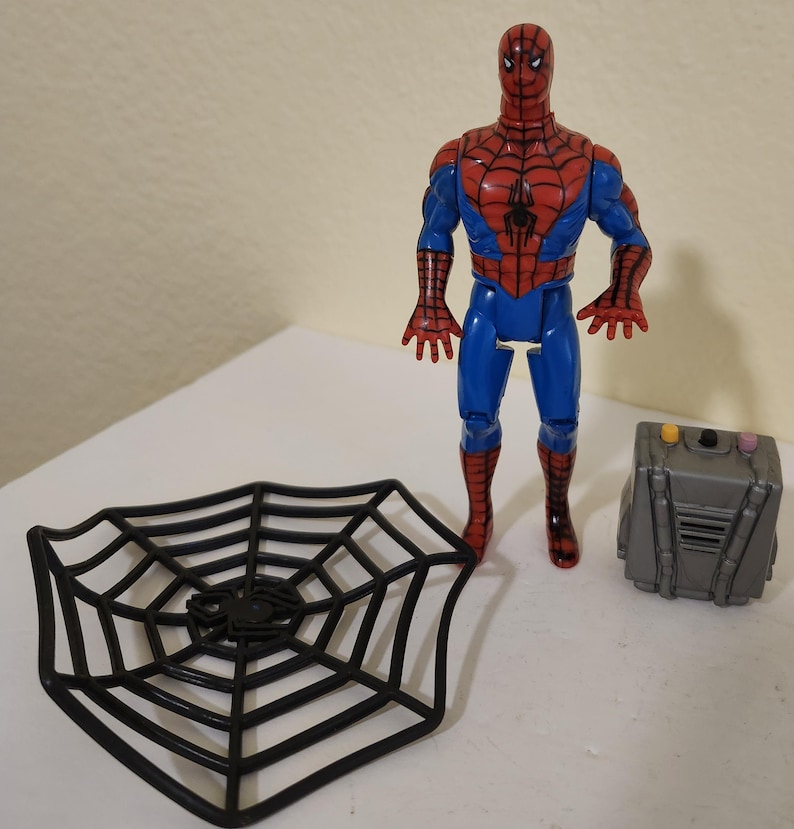 May include: A Spider-Man action figure in a classic red and blue suit, standing next to a black web-shaped accessory and a gray box. The figure has a detailed design, capturing the iconic superhero look.