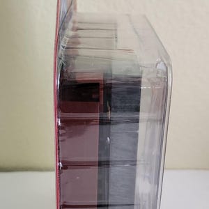 Peut inclure: Un emballage en plastique transparent contenant une figurine d'action rouge et noire. La figurine est partiellement visible &agrave; travers le plastique.