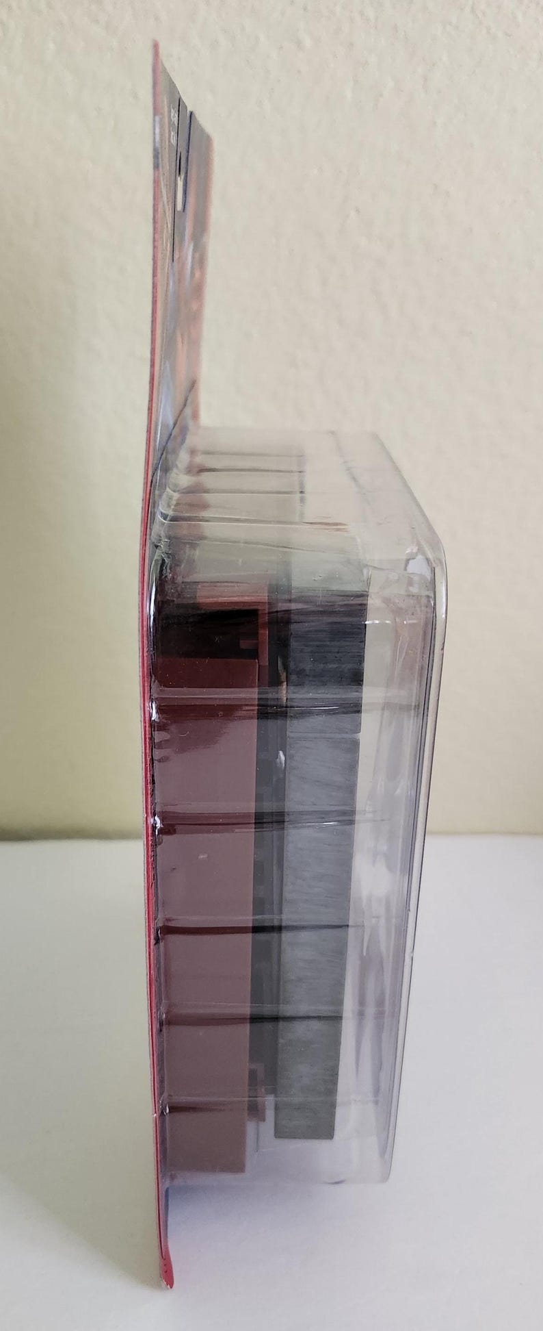 Peut inclure: Un emballage en plastique transparent contenant une figurine d'action rouge et noire. La figurine est partiellement visible &agrave; travers le plastique.
