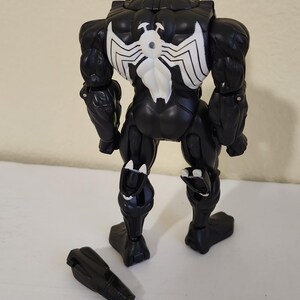 May include: Black Venom action figure with white spider emblem on the back. The figure has a detachable black and green accessory. The toy is posed on a white surface.