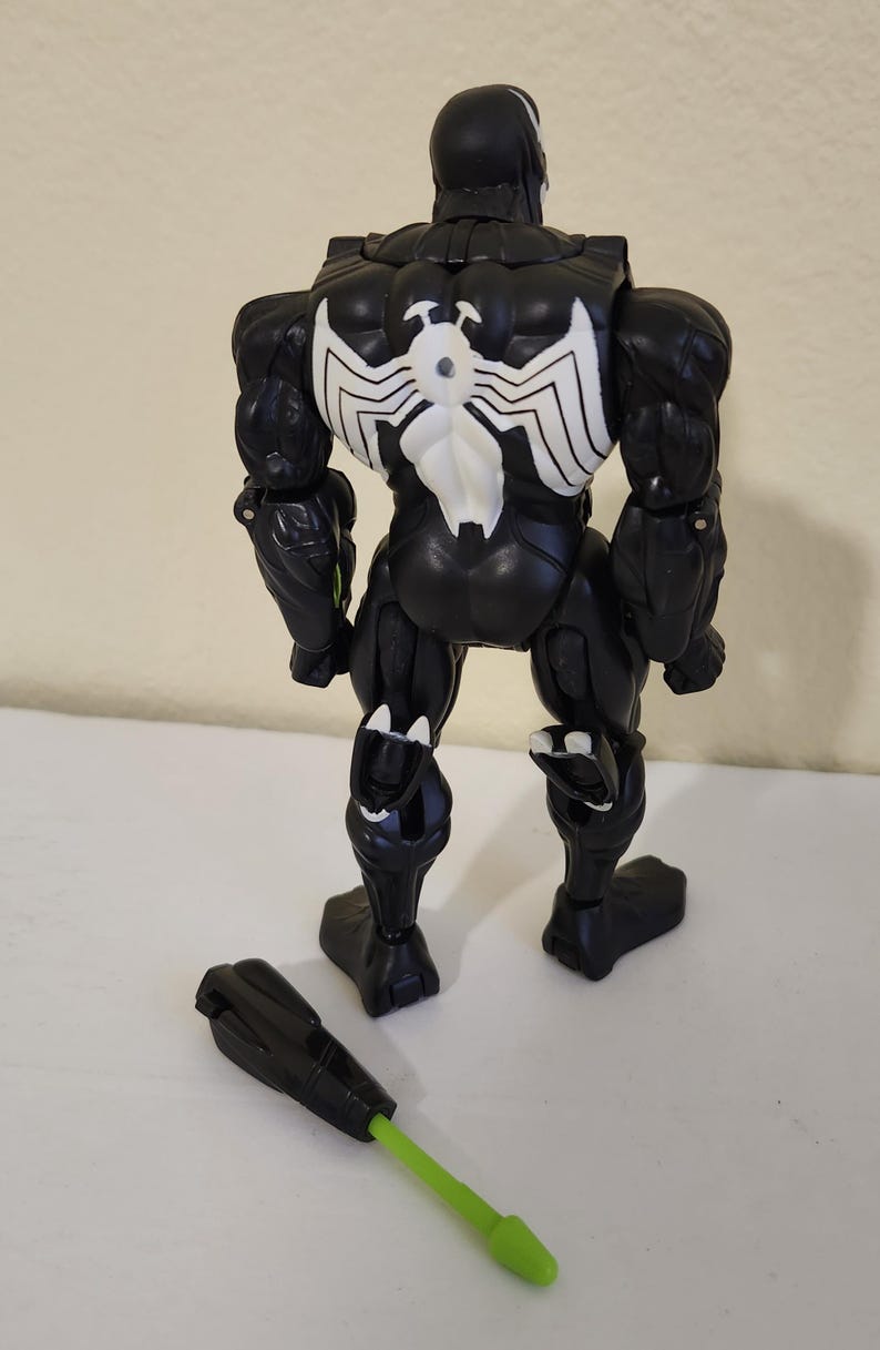 May include: Black Venom action figure with white spider emblem on the back. The figure has a detachable black and green accessory. The toy is posed on a white surface.