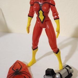 May include: A red action figure of Spider-Woman with black hair, a mask, and yellow accents. Accessories include a red spider-shaped shield and a black and silver weapon. The figure is standing on a white surface.