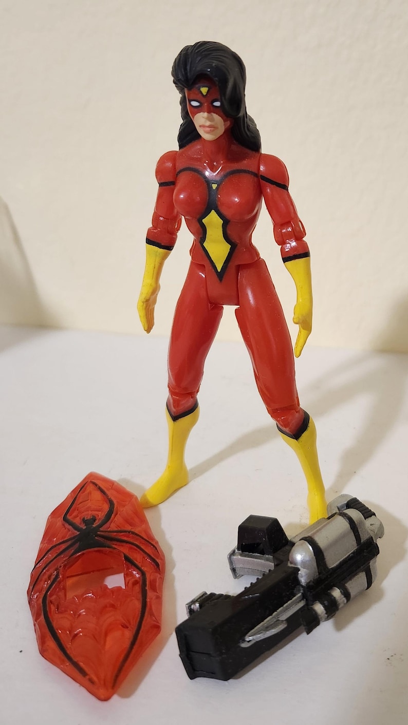 May include: A red action figure of Spider-Woman with black hair, a mask, and yellow accents. Accessories include a red spider-shaped shield and a black and silver weapon. The figure is standing on a white surface.