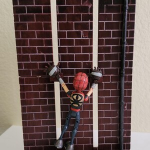 Peut inclure: Diorama miniature d'un mur de briques avec une figurine de super-h&eacute;ros. Le mur est rouge fonc&eacute; avec des lignes de mortier blanches, et la figurine est rouge, noire et beige. La figurine est en position d'escalade.