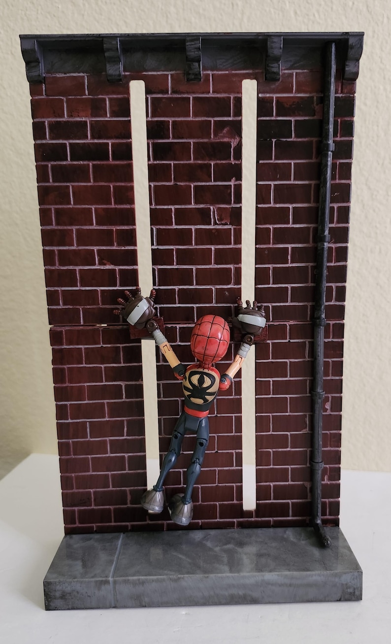 Peut inclure: Diorama miniature d'un mur de briques avec une figurine de super-h&eacute;ros. Le mur est rouge fonc&eacute; avec des lignes de mortier blanches, et la figurine est rouge, noire et beige. La figurine est en position d'escalade.