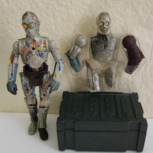 May include: A C-3PO action figure with a silver and gold finish, showcasing exposed wiring and mechanical details. The figure stands next to a clear plastic package containing additional parts and a dark gray storage box. A Star Wars collectible.