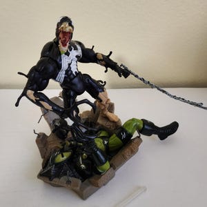 May include: A detailed action figure of Venom, a black and white superhero, in a dynamic pose, attacking a green and black figure. The figures are on a brown, rocky base, with a gray web-like strand extending from Venom's hand.