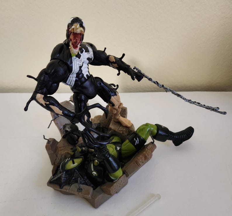 May include: A detailed action figure of Venom, a black and white superhero, in a dynamic pose, attacking a green and black figure. The figures are on a brown, rocky base, with a gray web-like strand extending from Venom's hand.