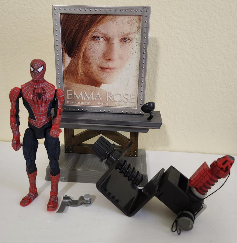 Może przedstawiać: Figurka Spider-Mana w czerwono-czarnym kostiumie stoi obok ekspozycji z oprawionym zdjęciem z napisem "EMMA ROSE PARFUMERIE PARIS NEW YORK". Obecne jest r&oacute;wnież czarno-czerwone urządzenie mechaniczne i inne akcesoria.