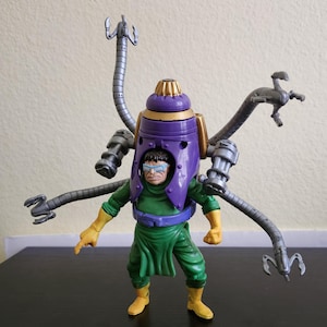 May include: A Dr. Octopus action figure. The figure is wearing a green outfit with yellow gloves and boots. A purple and gold helmet is on the figure's head, with four silver mechanical arms extending from the back.
