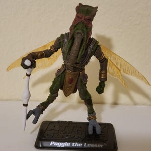 May include: A detailed action figure of Poggle the Lesser from the Star Wars universe. The figure is predominantly green and brown, with translucent yellow wings. It stands on a black base with the character's name printed on it. The figure is holding a white staff.