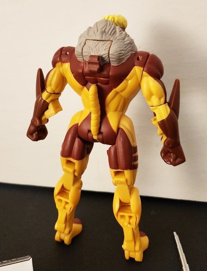 May include: A yellow and brown action figure of a character with a gray fur collar. The figure has a long tail and is posed with its arms at its sides.
