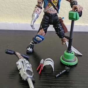May include: A detailed action figure of a mutated character with a blue vest, black shorts, and a "SMILE" tattoo. Accessories include a green weapon, a silver device, and other small parts. The figure is posed with a weapon.