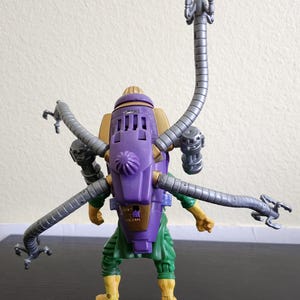 May include: A Dr. Octopus action figure, featuring a purple and gold backpack with articulated gray tentacles. The figure is wearing green pants, yellow boots, and has yellow arms.