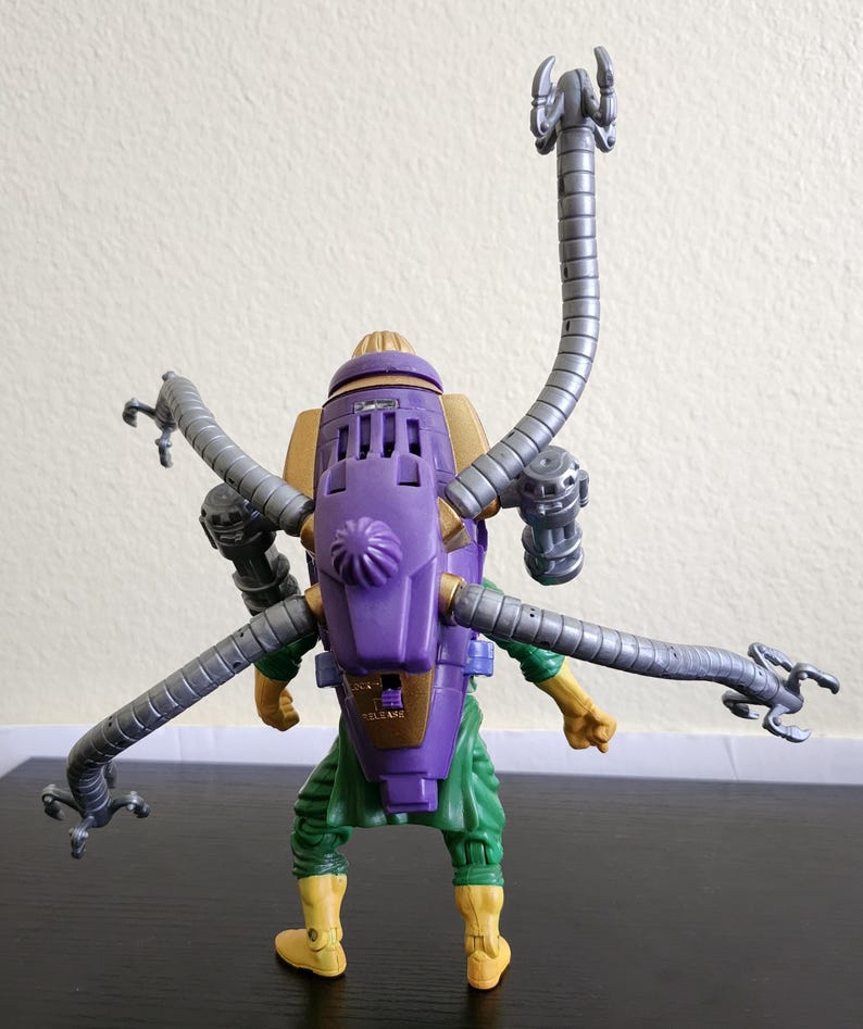 May include: A Dr. Octopus action figure, featuring a purple and gold backpack with articulated gray tentacles. The figure is wearing green pants, yellow boots, and has yellow arms.