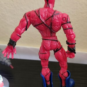 May include: A red action figure with black detailing and blue boots. The figure is posed with arms outstretched, showcasing the intricate design of the costume. The figure's back is visible, highlighting the sculpted muscles and lines.