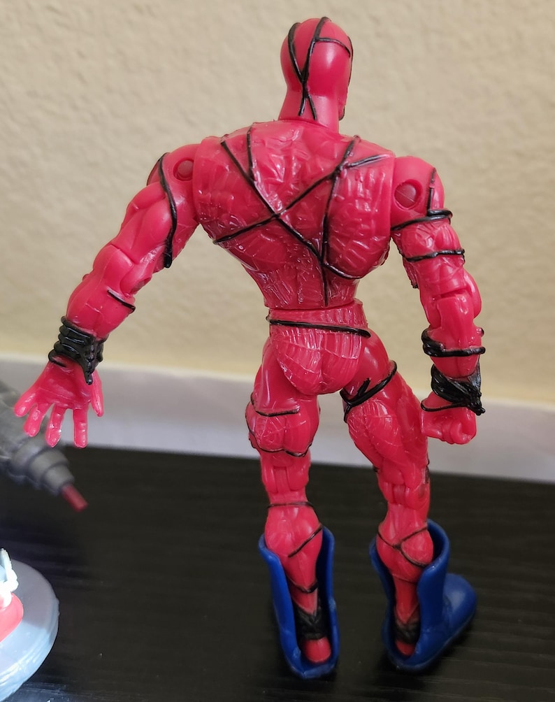 May include: A red action figure with black detailing and blue boots. The figure is posed with arms outstretched, showcasing the intricate design of the costume. The figure's back is visible, highlighting the sculpted muscles and lines.