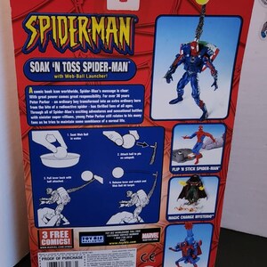 May include: A red, blue, and black Spider-Man action figure with a web-ball launcher. The figure is packaged in a cardboard box with a red background and the words "Soak 'N Toss Spider-Man" printed on it. The box also features images of other Spider-Man figures, including Flip 'N Stick Spider-Man, Magic Change Mysterio, and Aqua Blast Spider-Man.