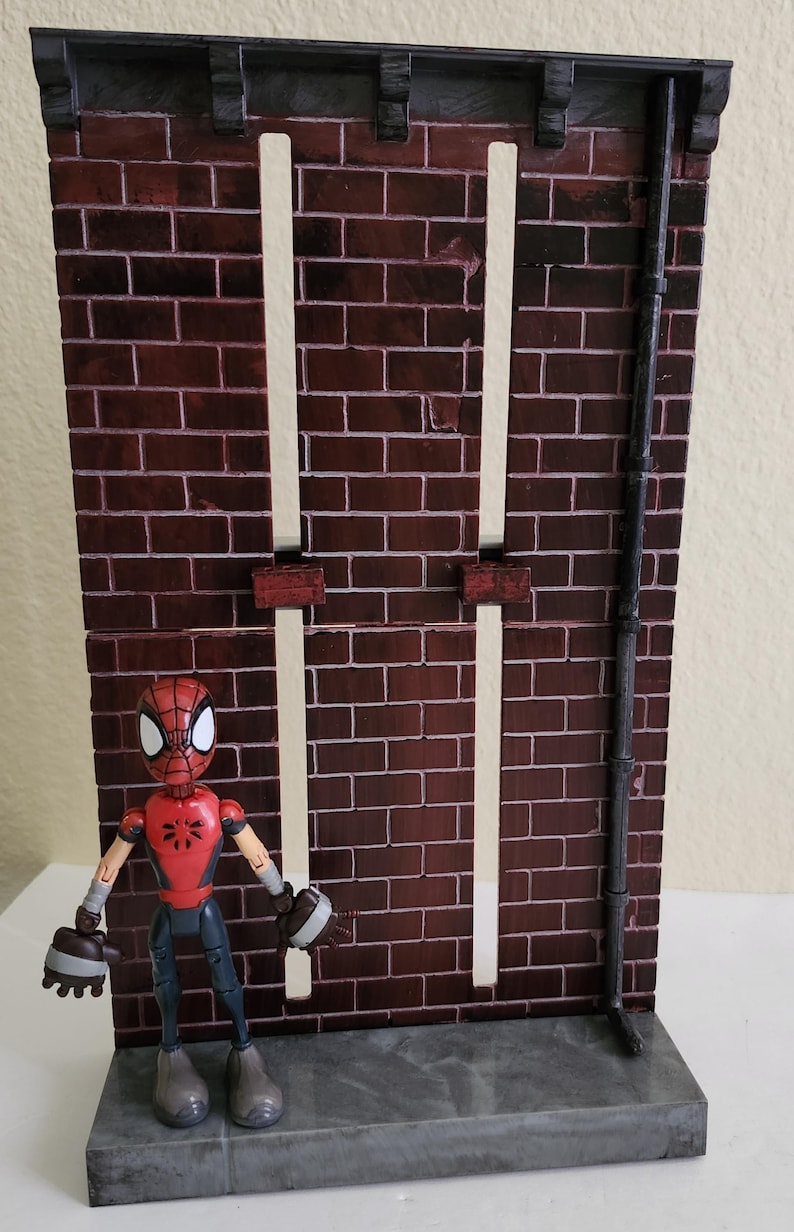 Peut inclure: Une figurine de Spider-Man se tient devant une toile de fond en briques. La figurine est rouge et noire, avec un masque et un costume d&eacute;taill&eacute;s. La toile de fond pr&eacute;sente un motif de briques sombres, avec des lignes verticales blanches et une goutti&egrave;re noire.