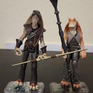 Könnte beinhalten: Zwei Star Wars-Actionfiguren der Gungan-Charaktere, Jar Jar Binks und ein weiterer Gungan. Die Figuren stehen auf transparenten Kunststoffsockeln. Eine Figur hält einen Stab, die andere eine speerartige Waffe. Die Figuren sind in Braun-, Beige- und Schwarztönen gehalten.