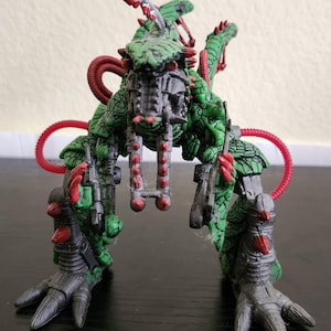 May include: A detailed action figure of a green and black creature with red accents. The figure has a complex design with multiple appendages, including clawed feet and a segmented body. The figure is posed on a dark surface.