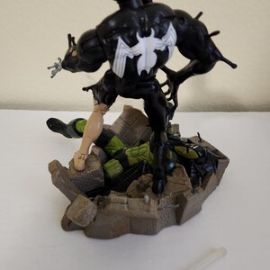 May include: A black Venom action figure with a white spider emblem on its chest, standing over a green and black figure on a brown base. A small, clear plastic accessory lies in the foreground.
