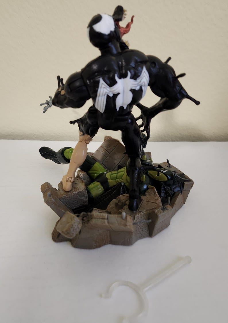 May include: A black Venom action figure with a white spider emblem on its chest, standing over a green and black figure on a brown base. A small, clear plastic accessory lies in the foreground.