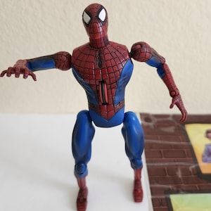 May include: A Spider-Man action figure in a dynamic pose. The figure features a red and blue costume with a web pattern on the torso and head. The figure's arms are outstretched, and the legs are bent, suggesting movement.