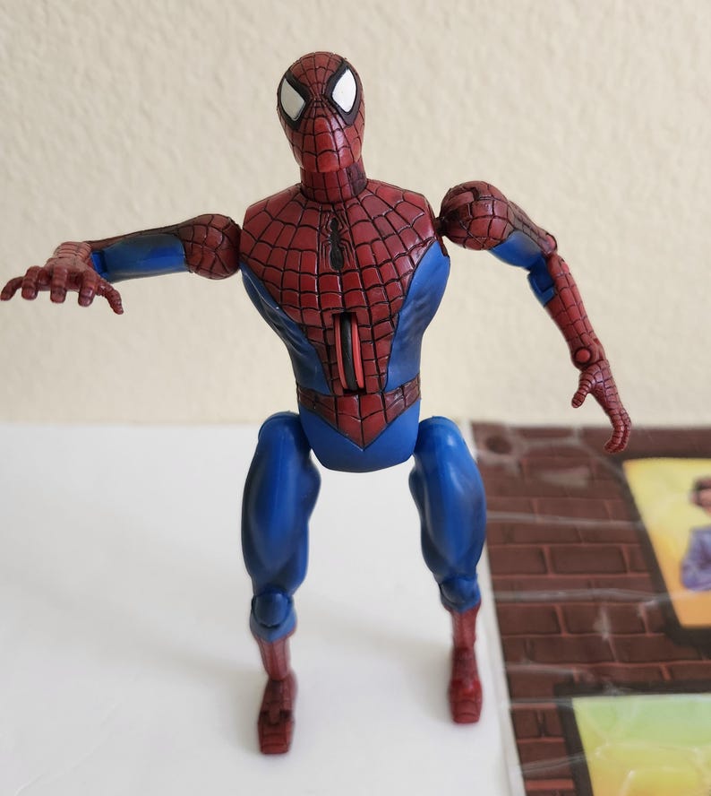 May include: A Spider-Man action figure in a dynamic pose. The figure features a red and blue costume with a web pattern on the torso and head. The figure's arms are outstretched, and the legs are bent, suggesting movement.