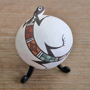 May include: A white ceramic decorative sphere with a black lizard design and geometric patterns in orange, brown, and green. The sphere sits on a black, three-legged stand. The lizard design wraps around the sphere.