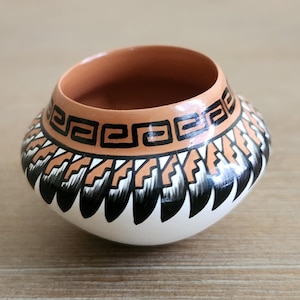 Peut inclure: Un petit bol en céramique fait à la main de forme arrondie. Le bol présente une base blanche, une bande de motifs géométriques noirs et beiges, et un intérieur couleur terre cuite. L'extérieur est orné de motifs complexes.
