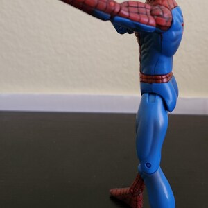 May include: A Spider-Man action figure in a dynamic pose, viewed from the side. The figure is predominantly blue and red, with a detailed web pattern on the red sections. The figure is striking a hand gesture.