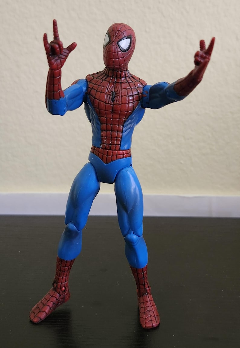 May include: A Spider-Man action figure in a dynamic pose, with arms raised and hands forming gestures. The figure features a red and blue costume with a spiderweb pattern on the red sections. The figure is standing on a dark surface.