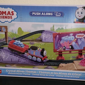 May include: Thomas & Friends Crystal Mines Thomas push-along train set. The blue train engine has a red caboose and is pulling a red ore car. The train is on a track with a sign that says "Thomas & Friends" and a pile of pink crystals.