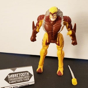May include: A brown and yellow action figure of Sabretooth, a Marvel Comics character, in his villain mode. The figure has a white fur collar and a yellow and brown tiger-like design. The figure is holding a silver sword. The figure is standing on a white surface. The figure is packaged with a card that says "Sabretooth Transforms into Sabretooth Tiger" and "Villain Mode Sabretooth".