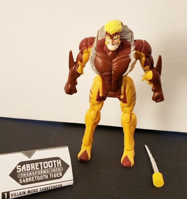 May include: A brown and yellow action figure of Sabretooth, a Marvel Comics character, in his villain mode. The figure has a white fur collar and a yellow and brown tiger-like design. The figure is holding a silver sword. The figure is standing on a white surface. The figure is packaged with a card that says "Sabretooth Transforms into Sabretooth Tiger" and "Villain Mode Sabretooth".