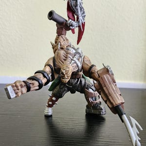 May include: A detailed action figure of a monstrous character with a large cannon and a smaller figure perched on its back. The main figure is brown and muscular, with a menacing expression. The smaller figure is silver and red.