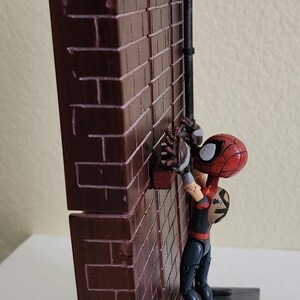 Peut inclure: Figurine de Spider-Man grimpant un mur de briques. La figurine porte un costume rouge et noir avec un masque distinctif. Le mur de briques est brun avec un toit noir et une conduite m&eacute;tallique.