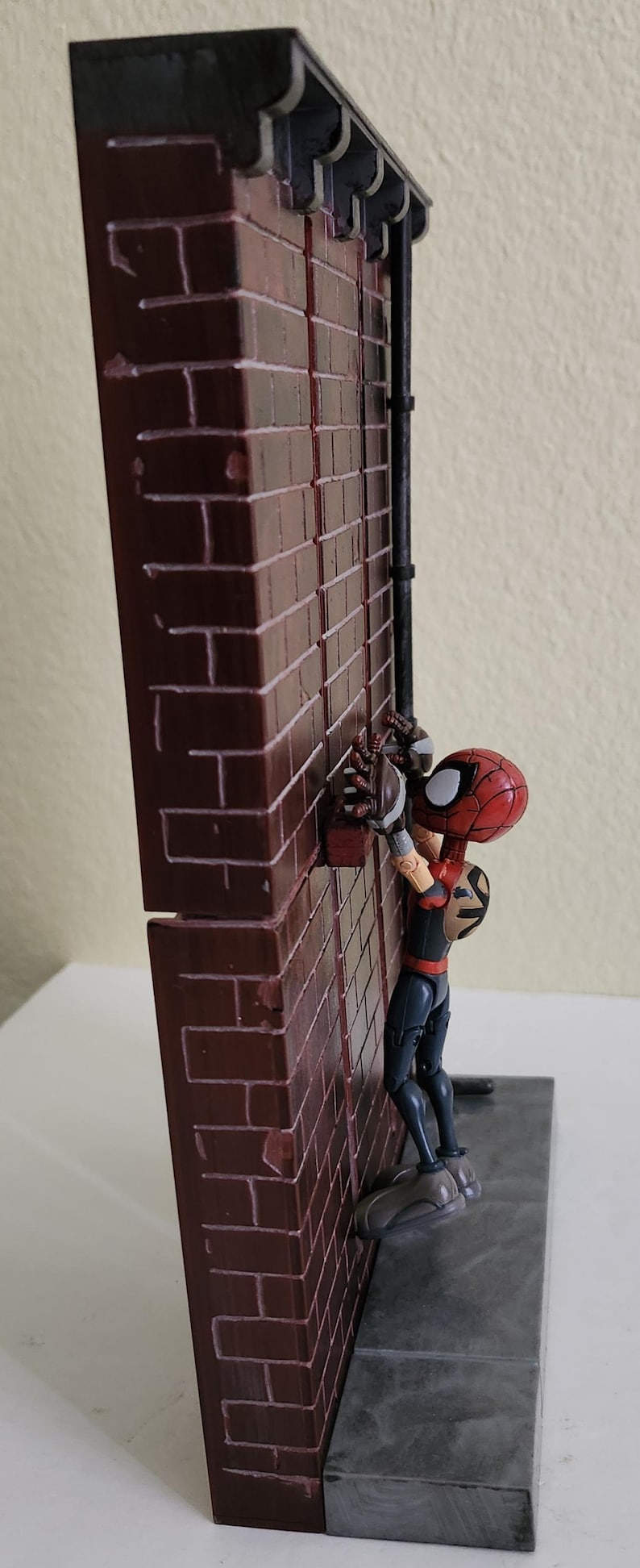 Peut inclure: Figurine de Spider-Man grimpant un mur de briques. La figurine porte un costume rouge et noir avec un masque distinctif. Le mur de briques est brun avec un toit noir et une conduite m&eacute;tallique.