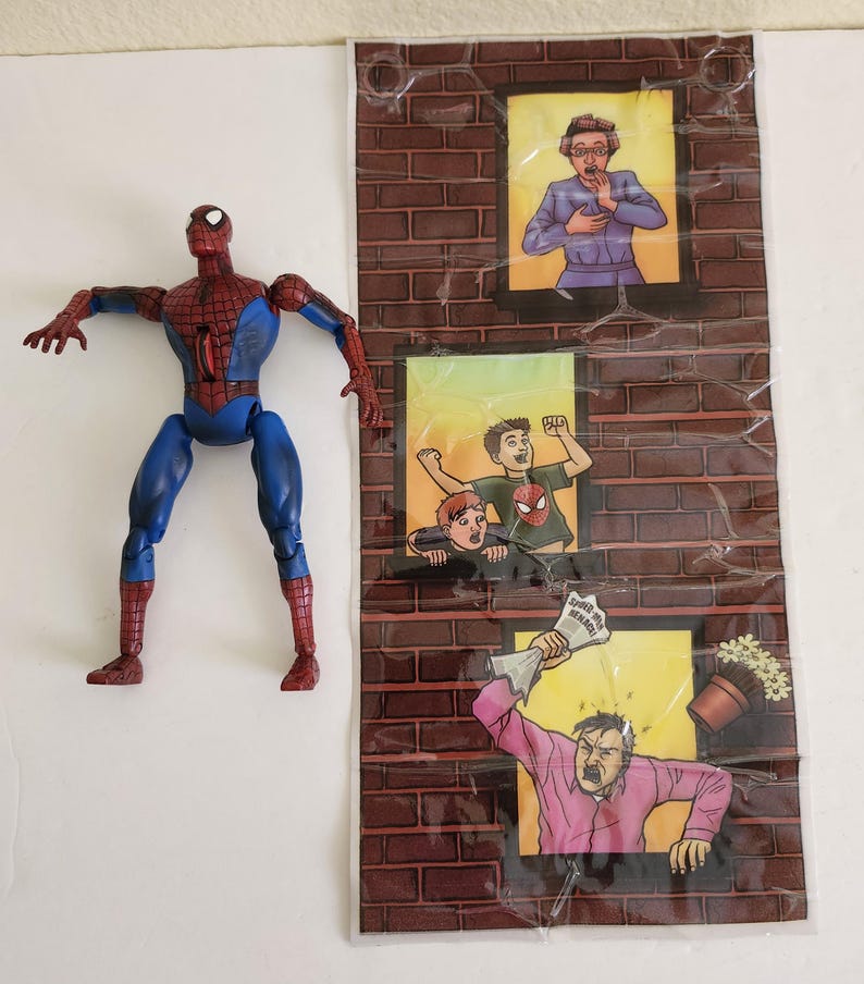 May include: A Spiderman action figure with a red and blue costume stands next to a brick wall backdrop. The backdrop features three windows with cartoon illustrations of people reacting to something. The text "Spider-Man Returns" is visible.